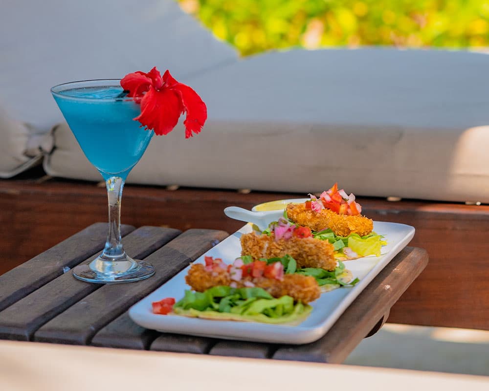 A blue cocktail with a red flower garnish alongside a plate of three crispy fish tacos topped with lettuce and diced tomatoes.