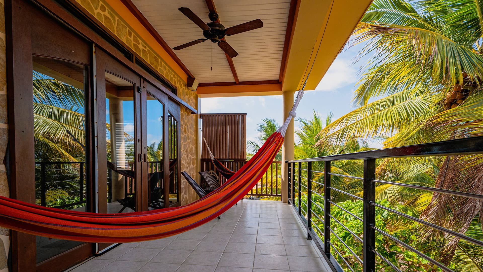 A colorful hammock hangs on a balcony surrounded by palm trees and bright sky.